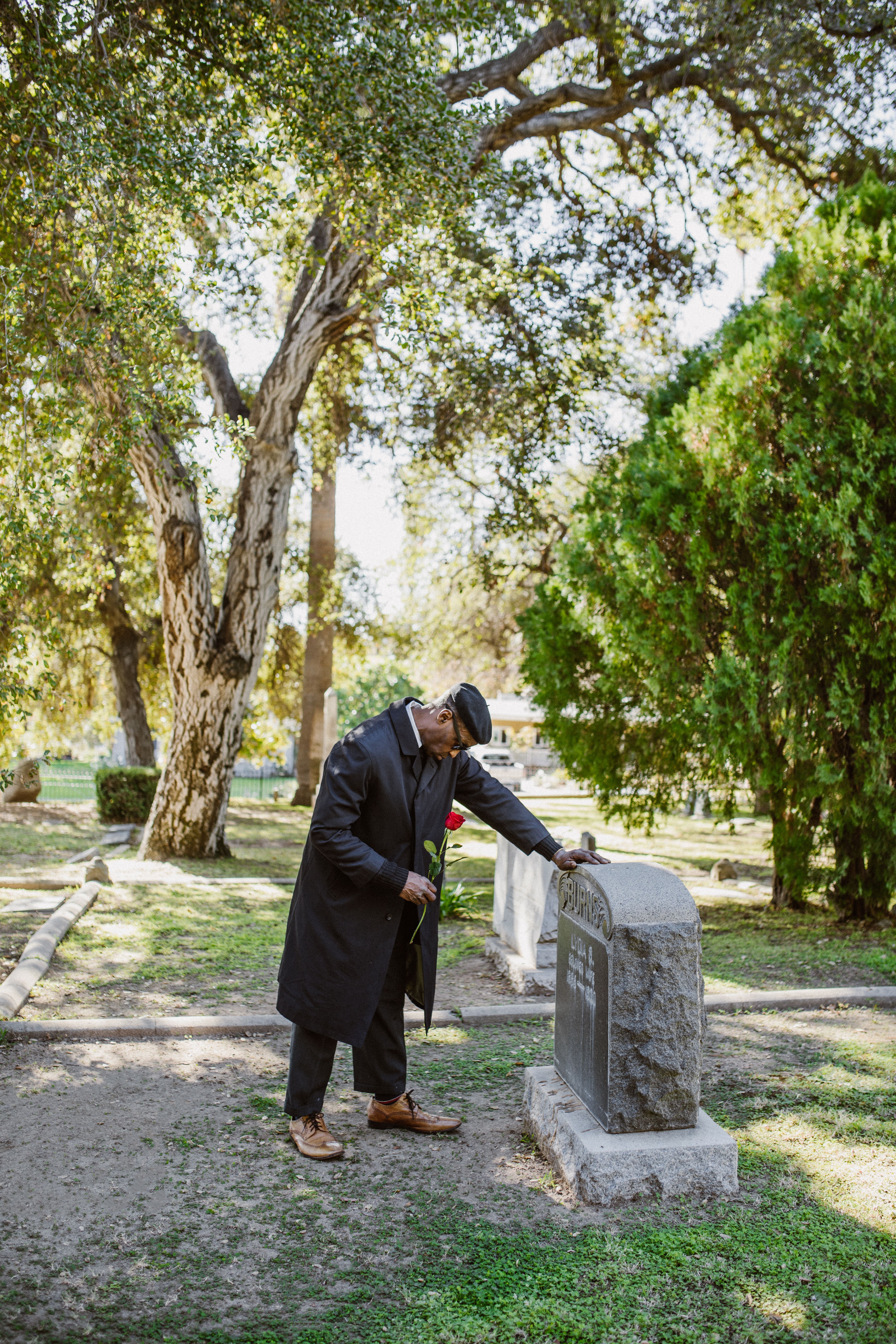 A man holding a rose in a cemetery, grieving and standing by a gravestone.