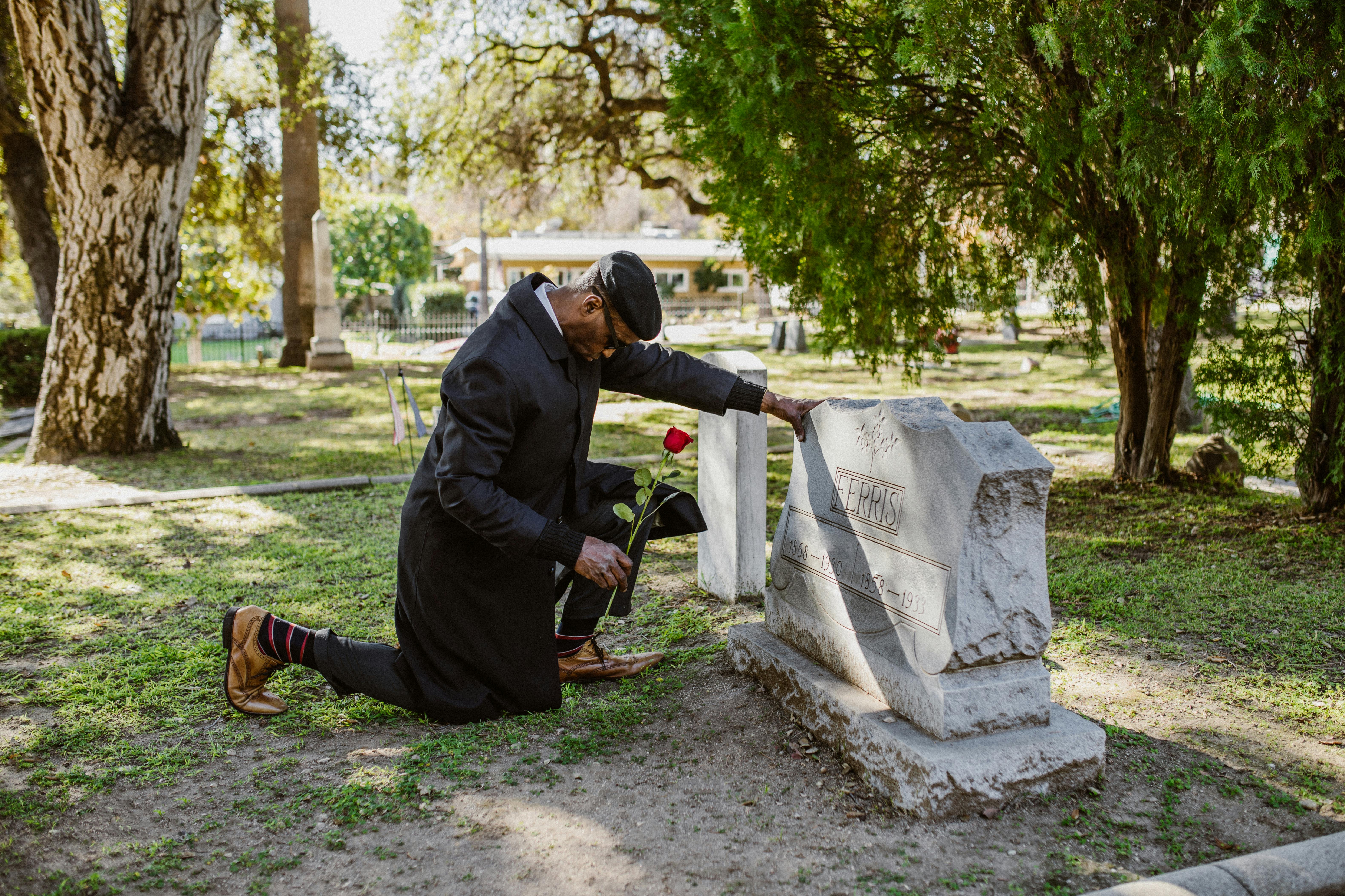 A man kneels at a gravestone, holding a red rose, mourning in a peaceful cemetery.