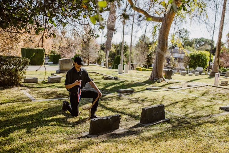 A Man In Black Clothes Kneeling On The Graveyard Holding A Medal