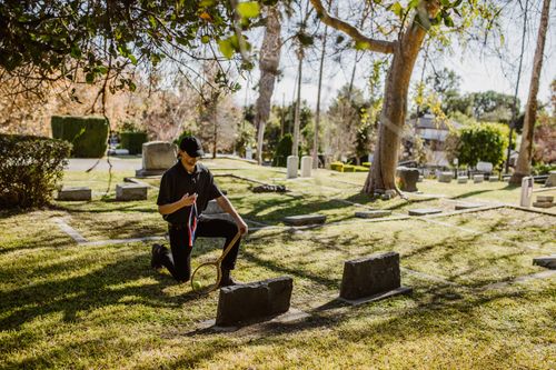 Free A man kneeling with a memento in a tranquil cemetery surrounded by tombstones and trees. Stock Photo
