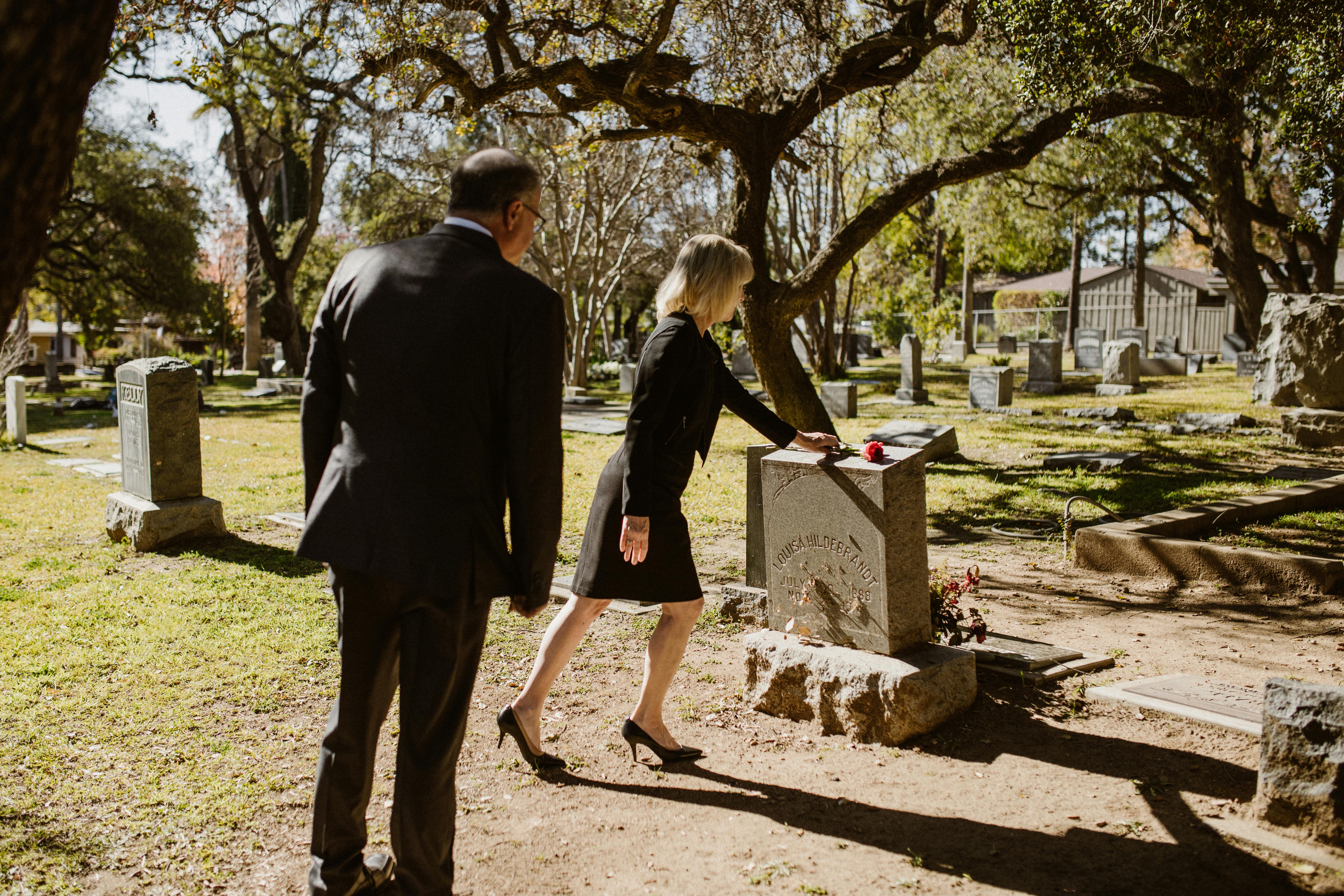 A somber moment as a couple places a rose on a gravestone in a peaceful cemetery.