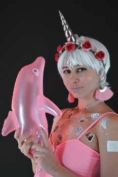 Young female model with creative makeup and stickers on skin wearing wig with horn holding inflated dolphin against black background