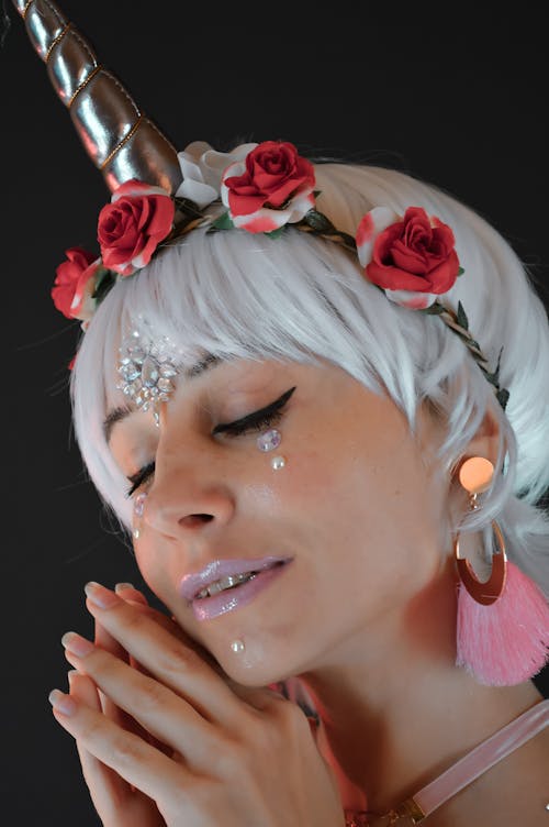Serene female with creative makeup and wig with unicorn horn closing eyes against black background