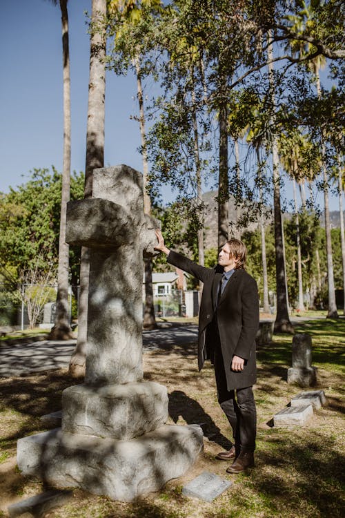 
A Man in a Black Coat Touching a Stone Crucifix