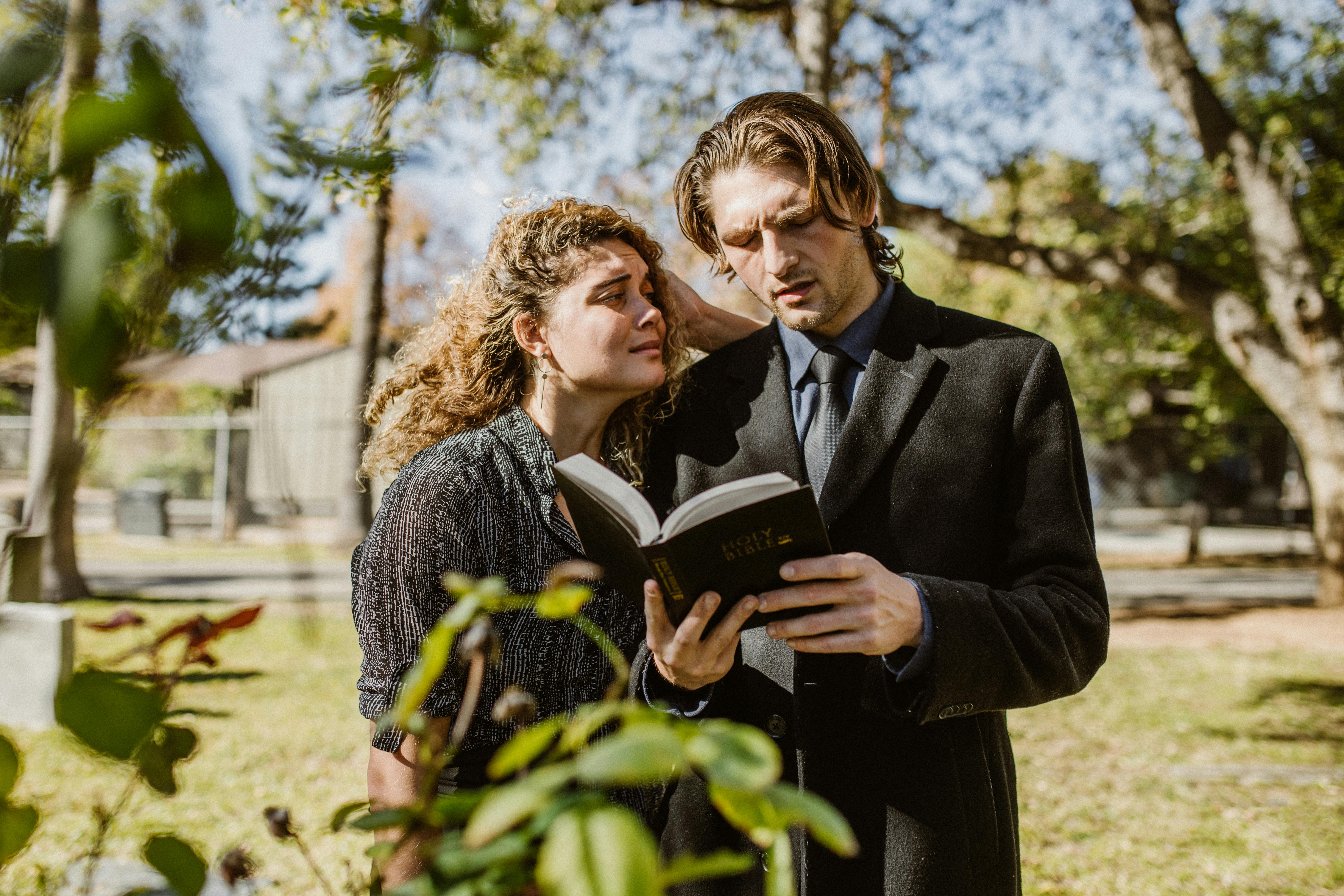 man in black suit jacket reading a bible beside a grieving woman in black coat