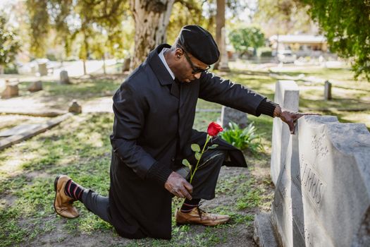 A man kneels at a gravestone in a cemetery holding a red rose, reflecting loss and remembrance.