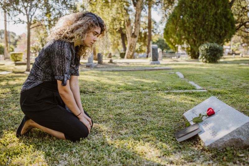 Mulher depositando uma rosa vermelha sobre um túmulo em um jardim de cemitério, homenagem a quem faleceu