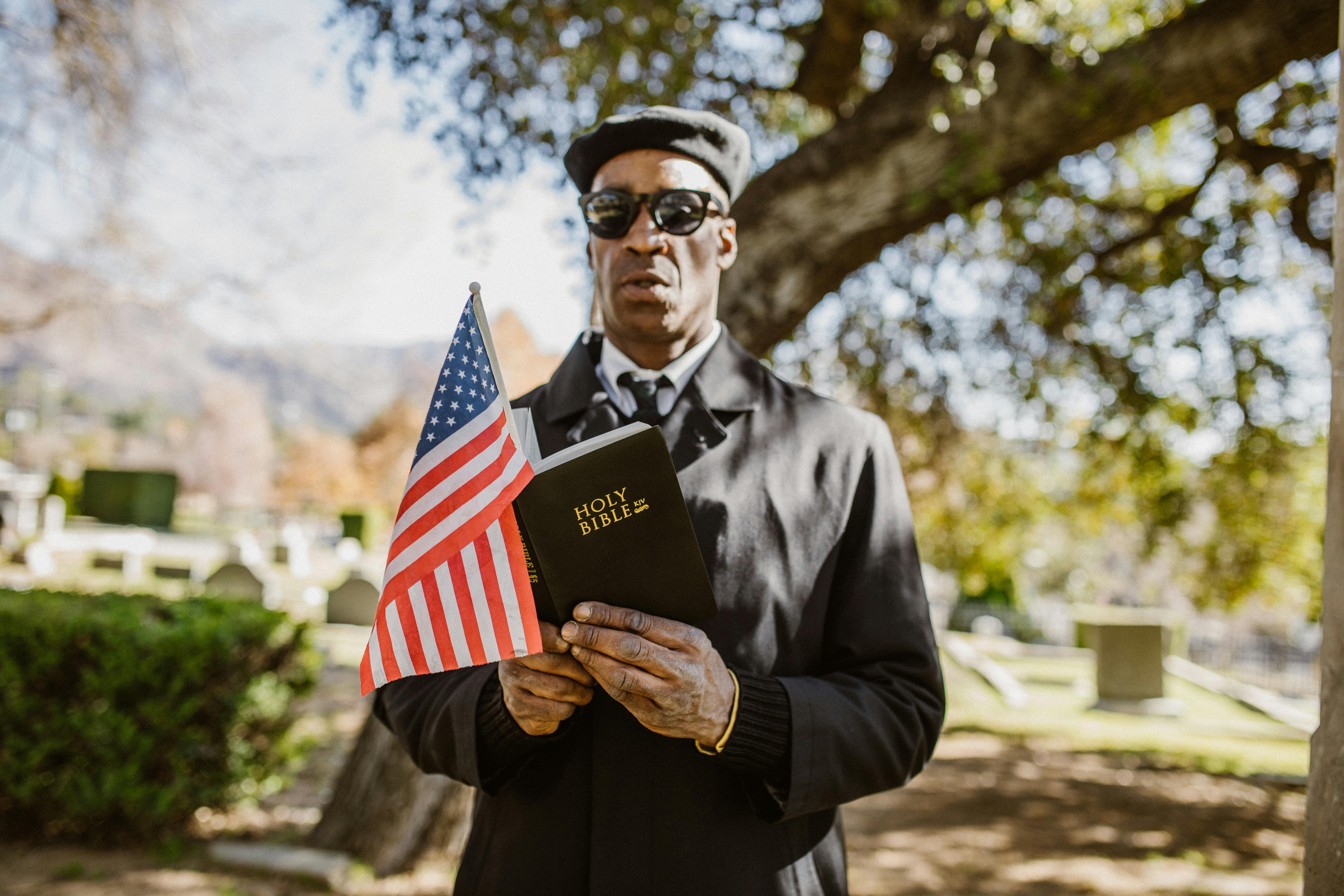 a man in a black coat reading the bible while holding an american flag