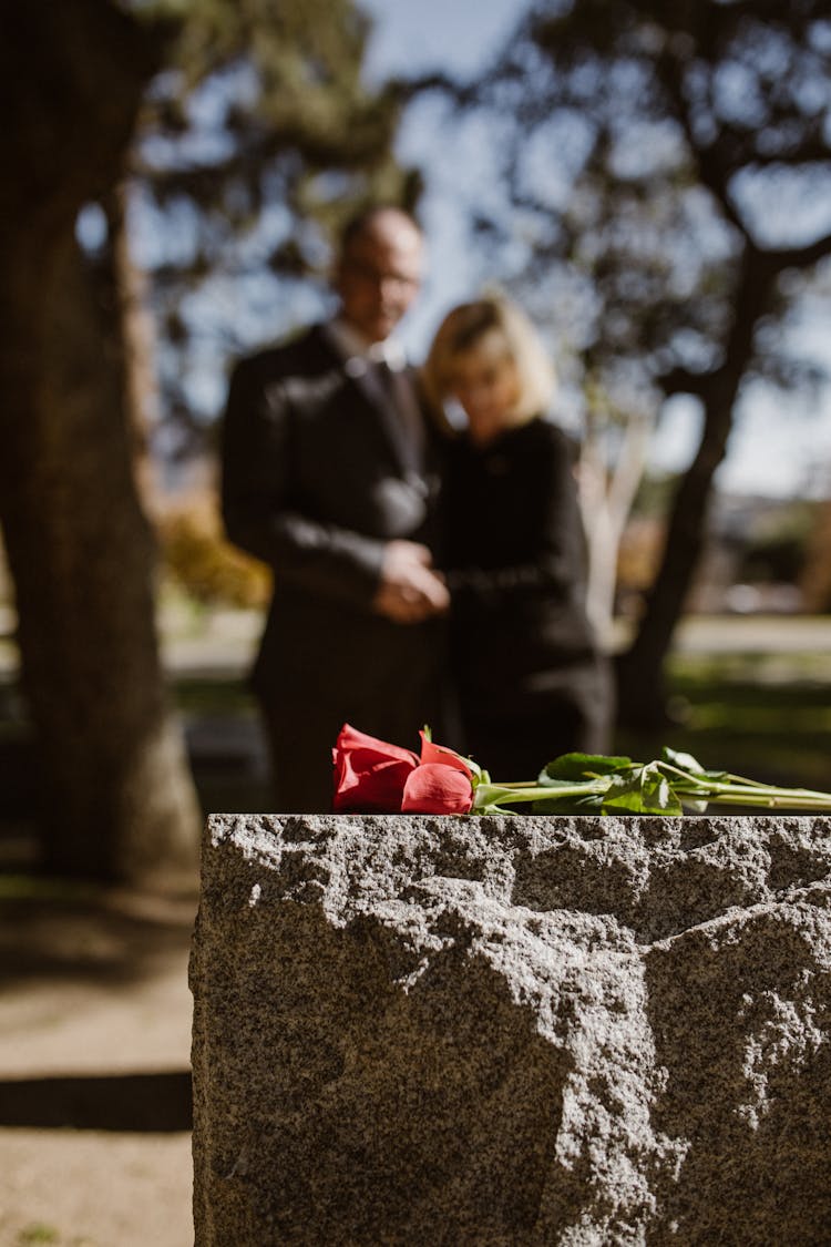 Man And Woman Standing On Grave With Flower On Tombstone