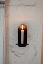 Burning wax candle in metal holder hanging on rough whitewashed wall in aged building