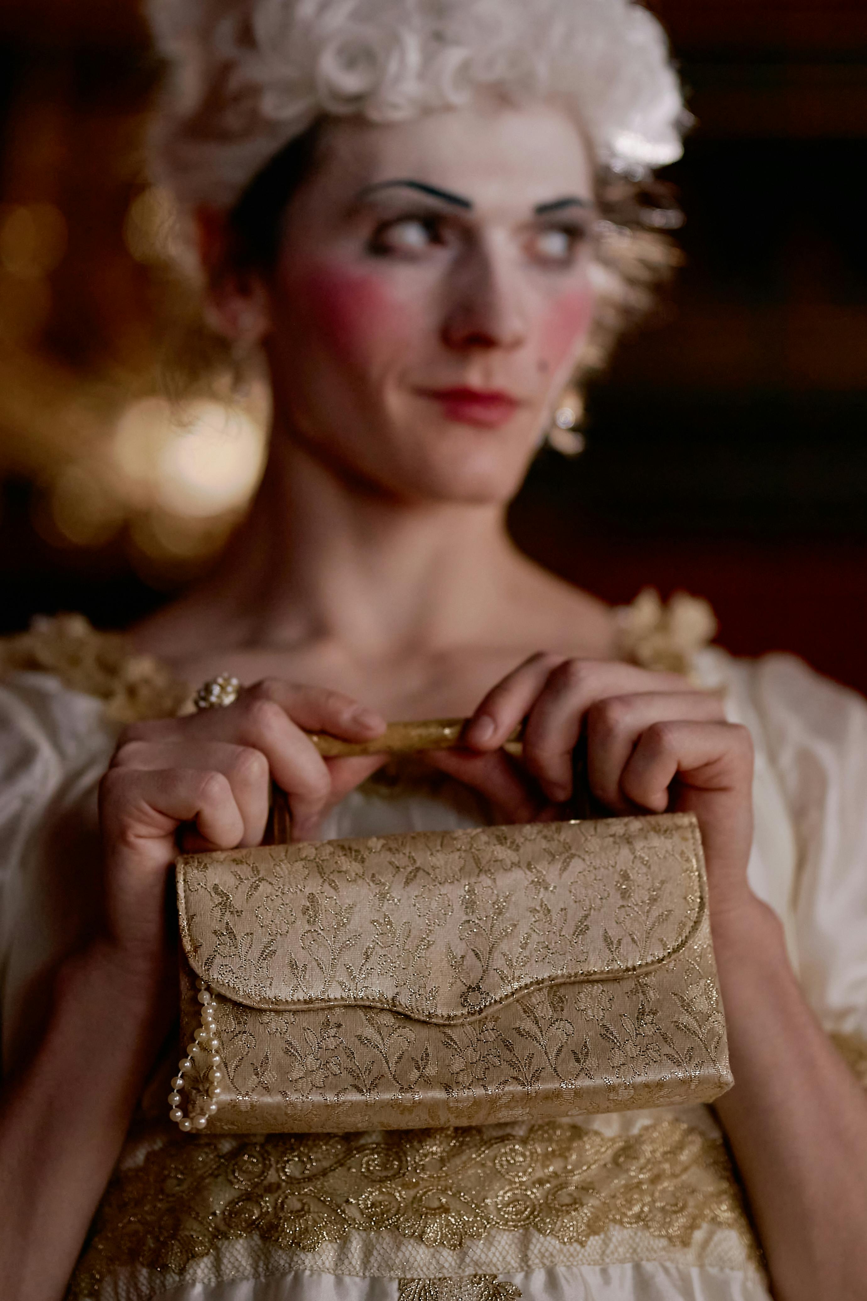 Free A cross-dresser elegantly holds a golden purse in a baroque-style setting, showcasing vintage fashion. Stock Photo