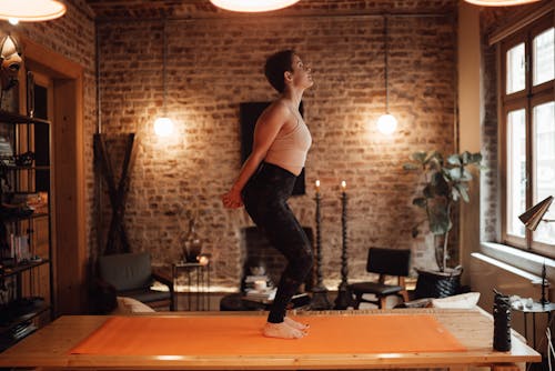 Free Side view full body of slim barefoot lady in activewear standing on yoga mat on wooden platform while exercising with hands behind back in light apartment Stock Photo