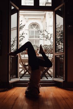 Full length side view of faceless slim barefoot person in casual clothes standing on head in apartment on wooden floor near windows with balcony in daylight