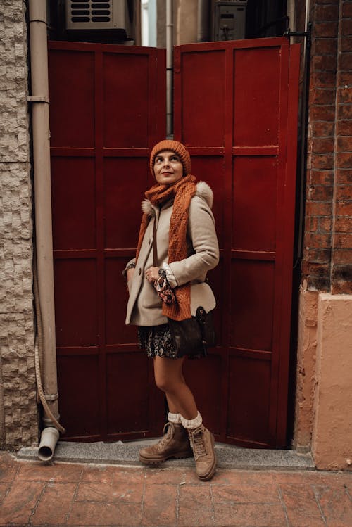 Free Full body of young female in casual clothes and knitted hat standing near metal door of aged building and looking up while walking in city Stock Photo