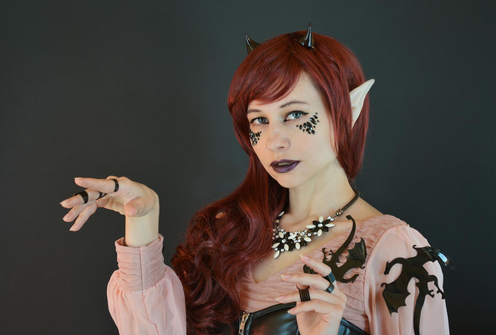 Creative young female model with long red hair and artificial ears and horns in elf costume looking at camera against black background