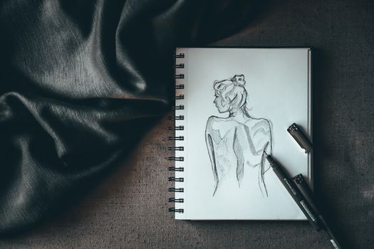 Top view composition of spiral notebook with sketch of undressed female placed on dark surface near liners and black silk fabric