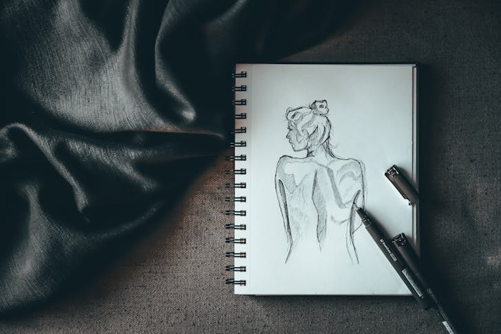 Top view composition of spiral notebook with sketch of undressed female placed on dark surface near liners and black silk fabric