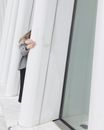 Crop unrecognizable female in casual clothes standing behind white pillar on floor near glass window in light building