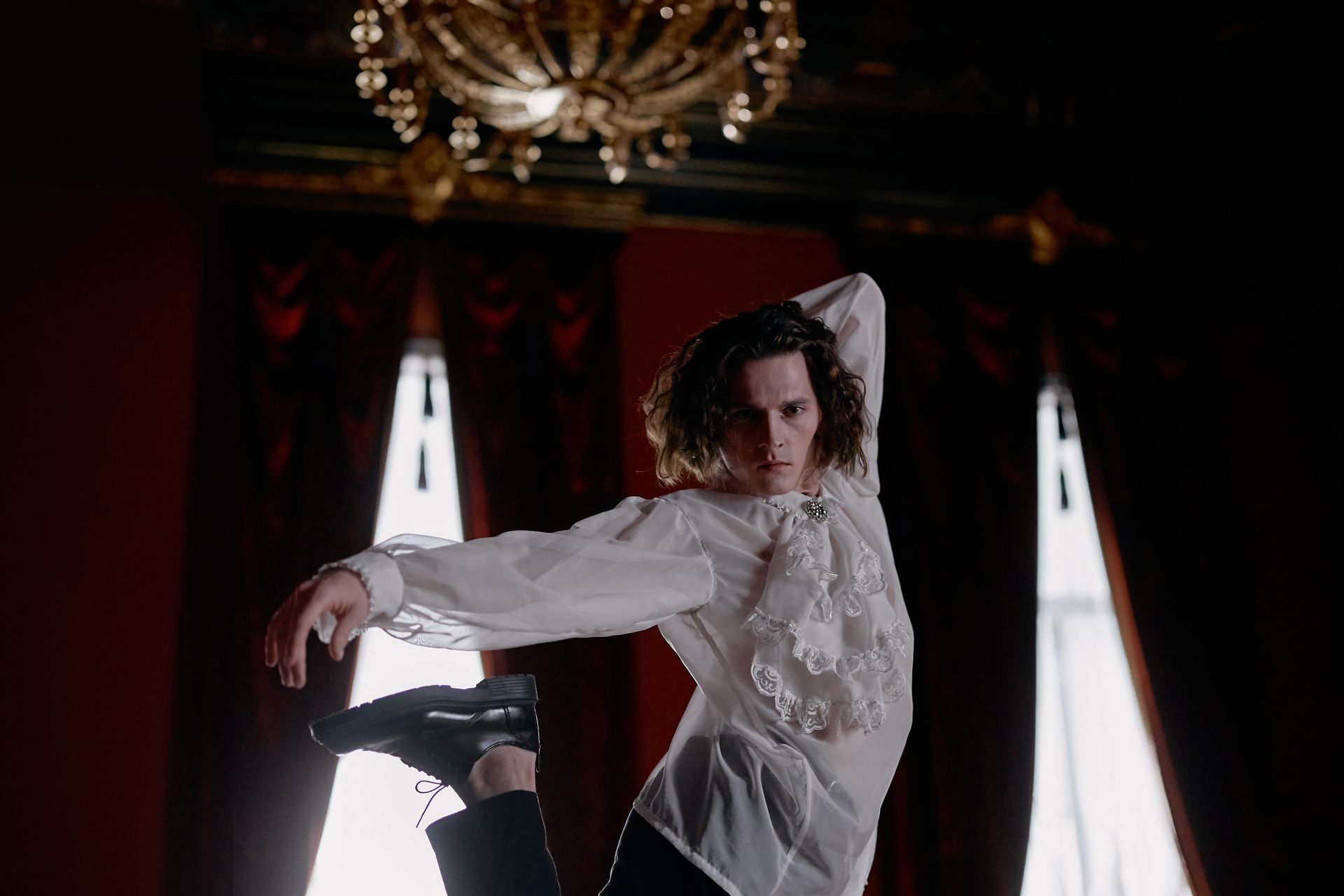 An adult male dancer gracefully poses under an ornate chandelier indoors.