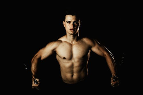 Young sporty male with naked torso exercising with cable crossover machine and looking at camera on black background