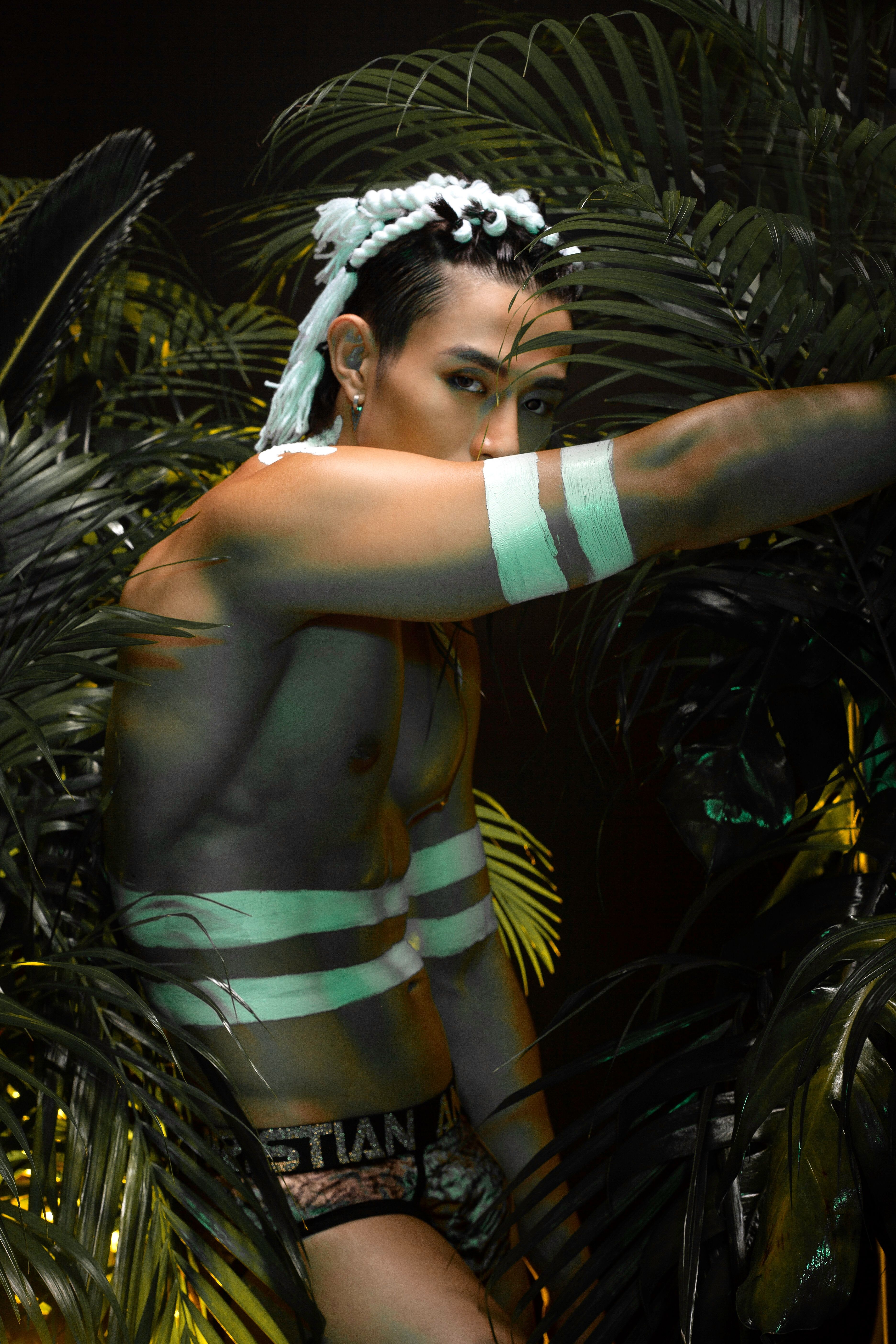 Striking image of a man with body paint and stylish hair amidst lush jungle leaves, showcasing modern fashion.