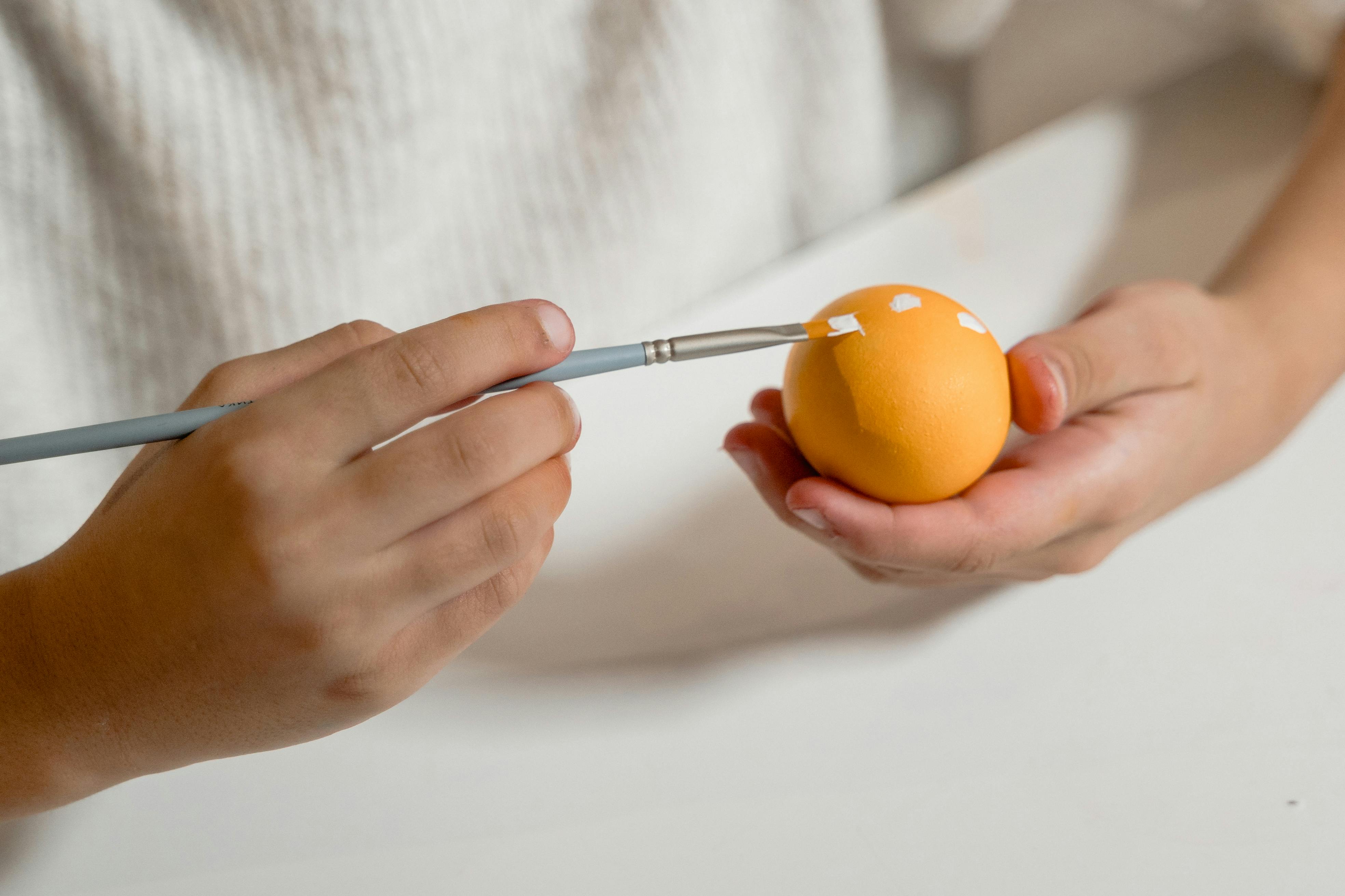 Creative close-up of hands holding and painting a yellow Easter egg with a paintbrush.