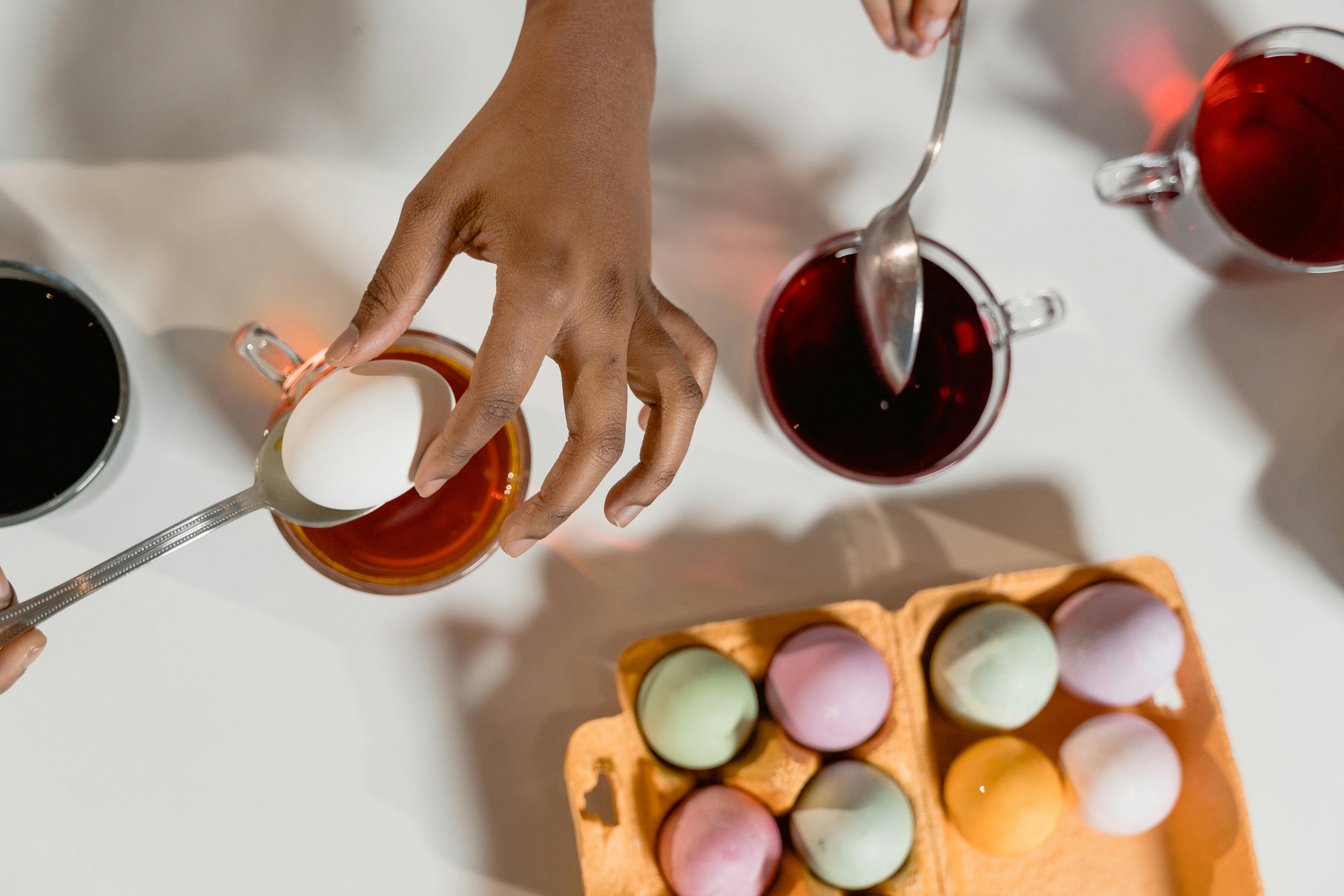 Hands dyeing Easter eggs using various colors in a kitchen setting.