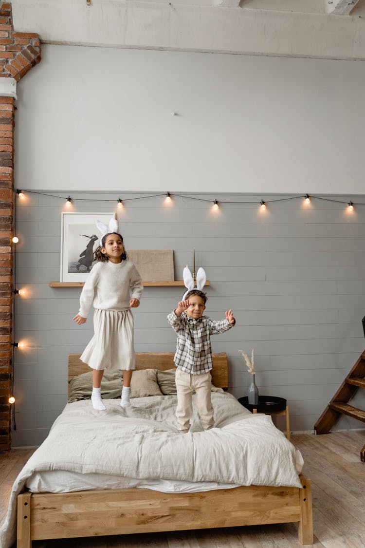 A Girl Jumping On Bed Beside A Boy Wearing Bunny Ears Headband