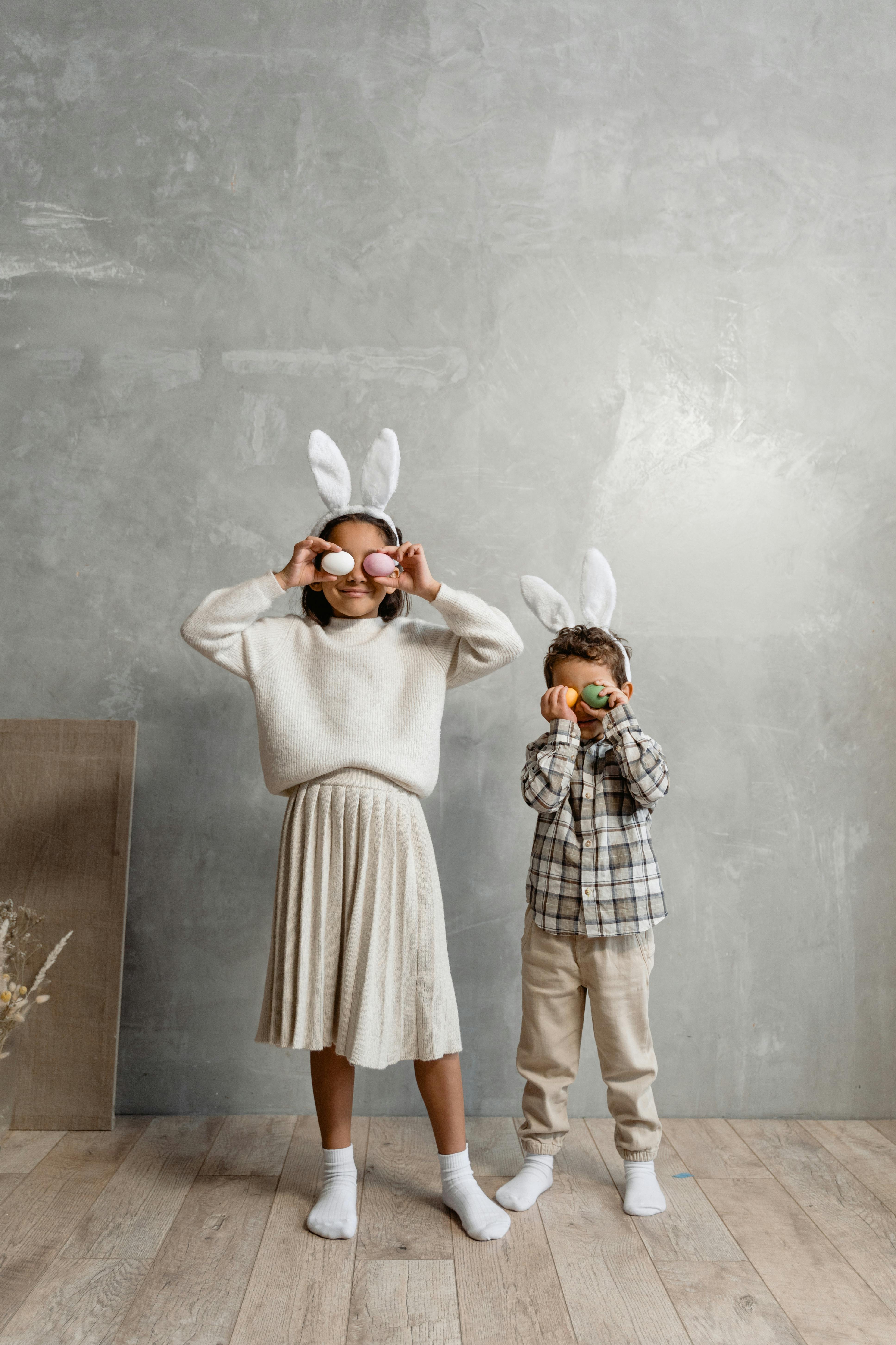 a girl and a boy wearing bunny ears headband covering their eyes with eggs