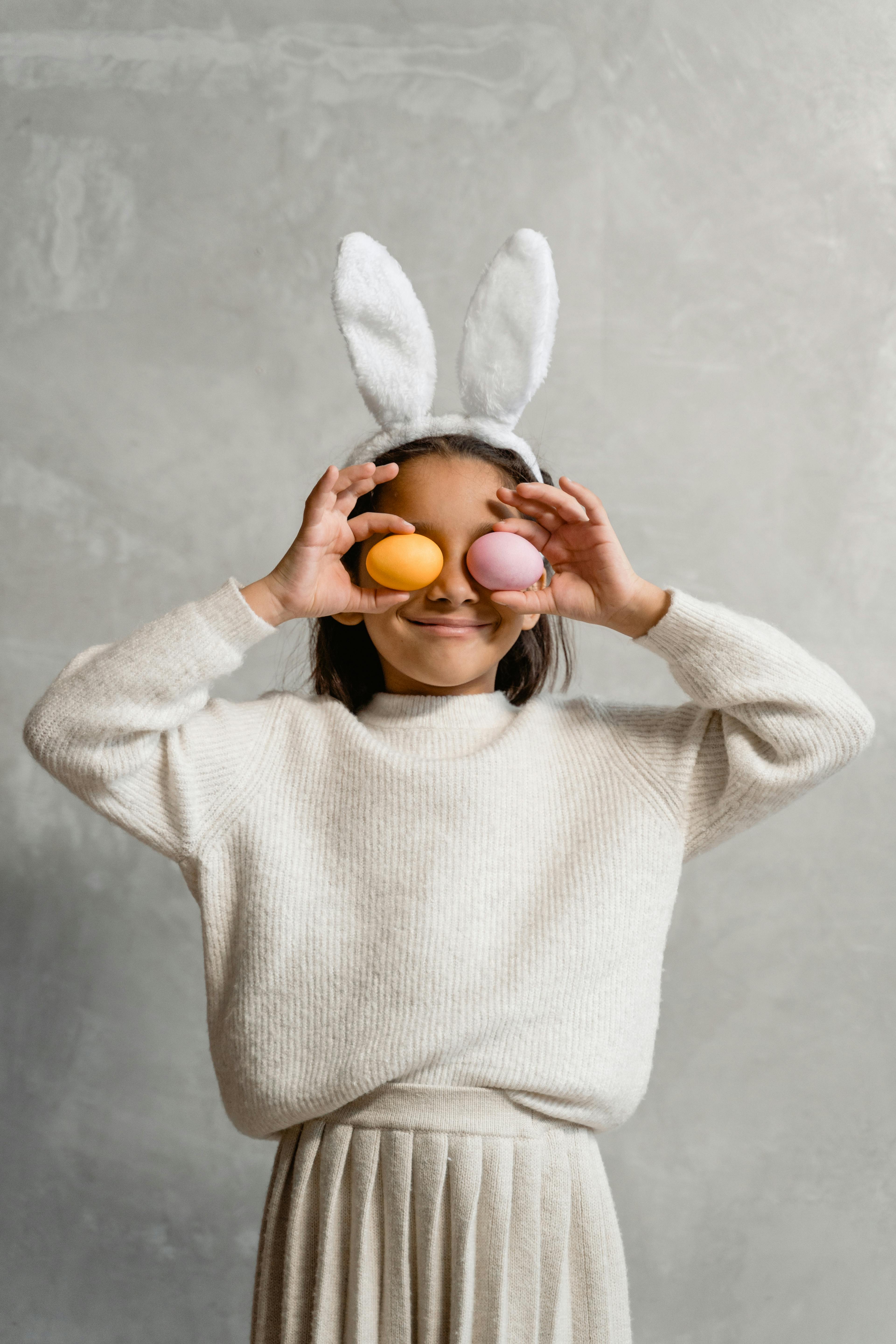 Adorable child with bunny ears playfully holding colored Easter eggs over eyes.