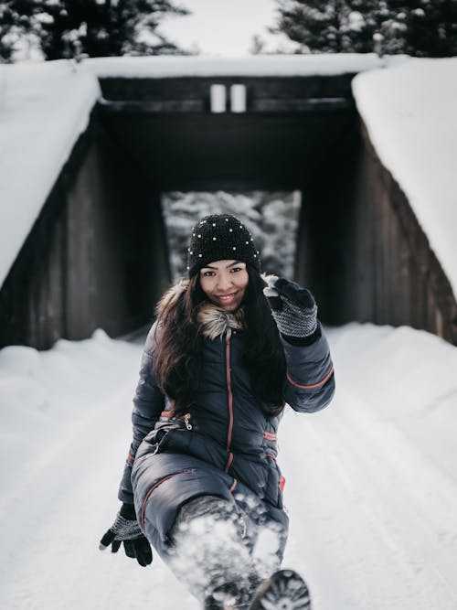 Cheerful ethnic woman kicking snow in winter park