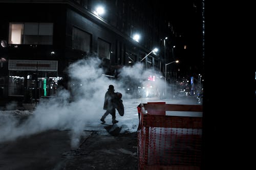 Side view of distant anonymous person crossing asphalt roadway on street with steam and residential buildings at night time in city