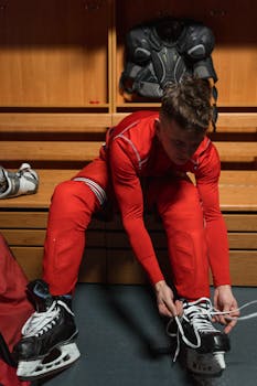 An ice hockey player in a red uniform lacing skates in a locker room.