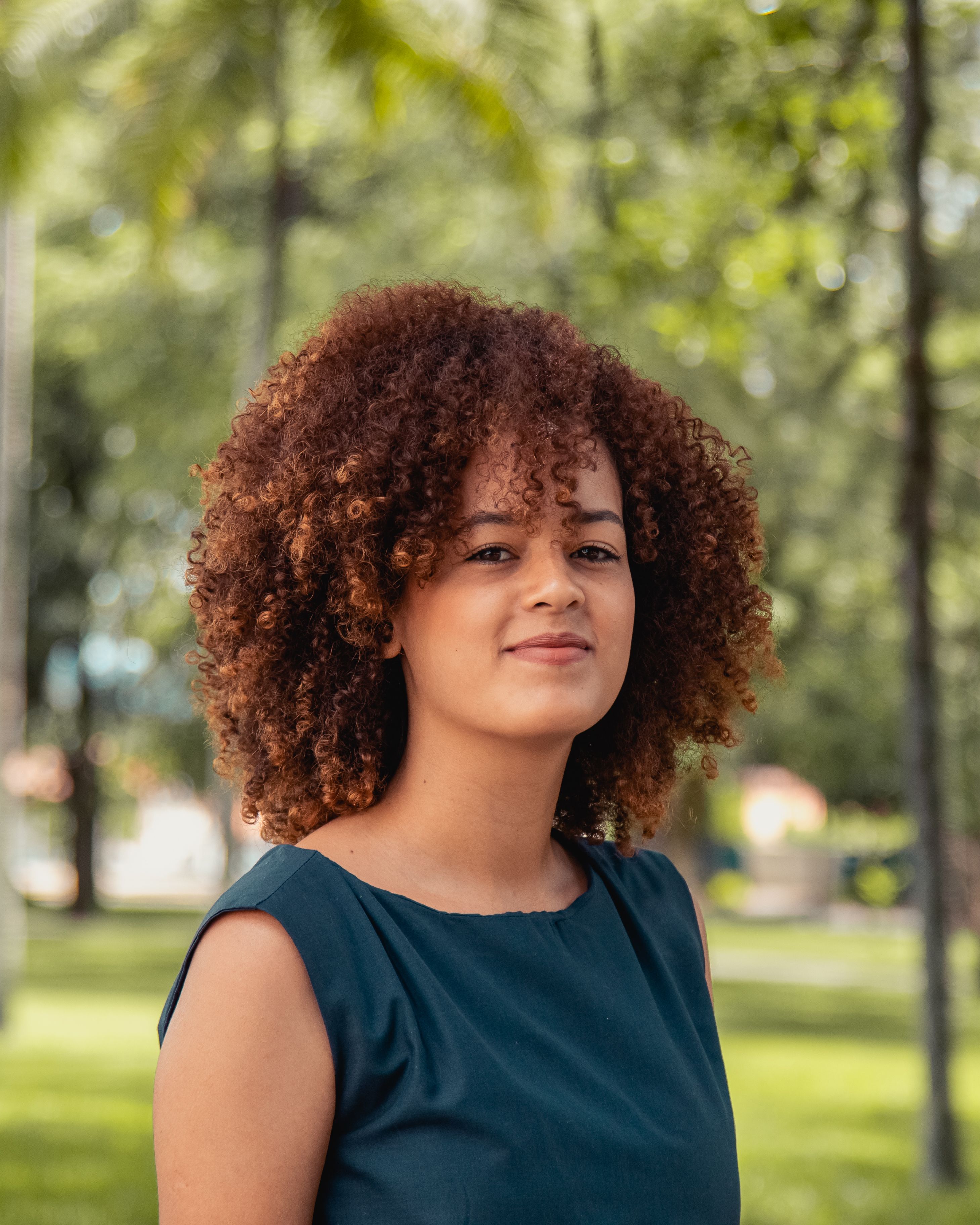 Smiling woman with curly hair standing outside in a lush green park. Natural light highlights her joyful expression.