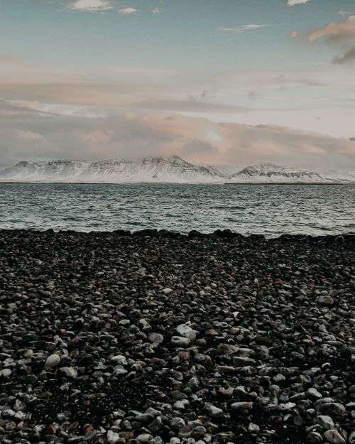 Free Shore with pebbles near rippling sea water against cloudy sky and mountain range covered with snow in cold winter day Stock Photo