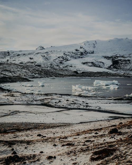 Calm lake with ice chunks on water surface surrounded with mountains covered with snow on cold winter day in nature