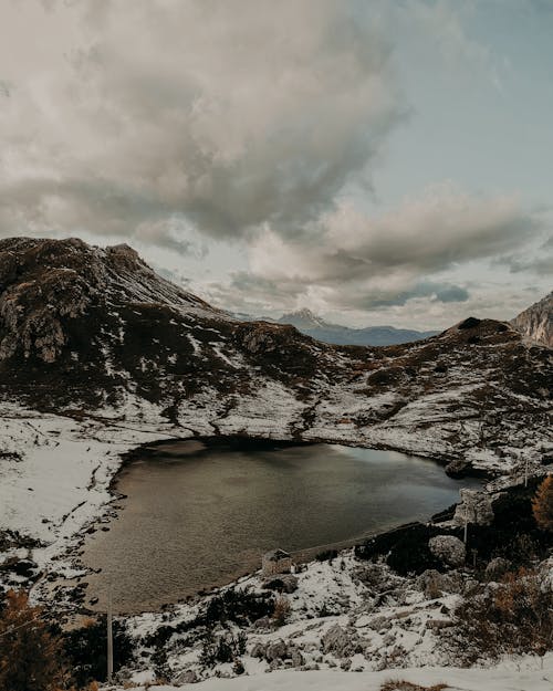 Small lake with calm frozen water surrounded by snowy rocky formations against cloudy sky in nature on cold winter day