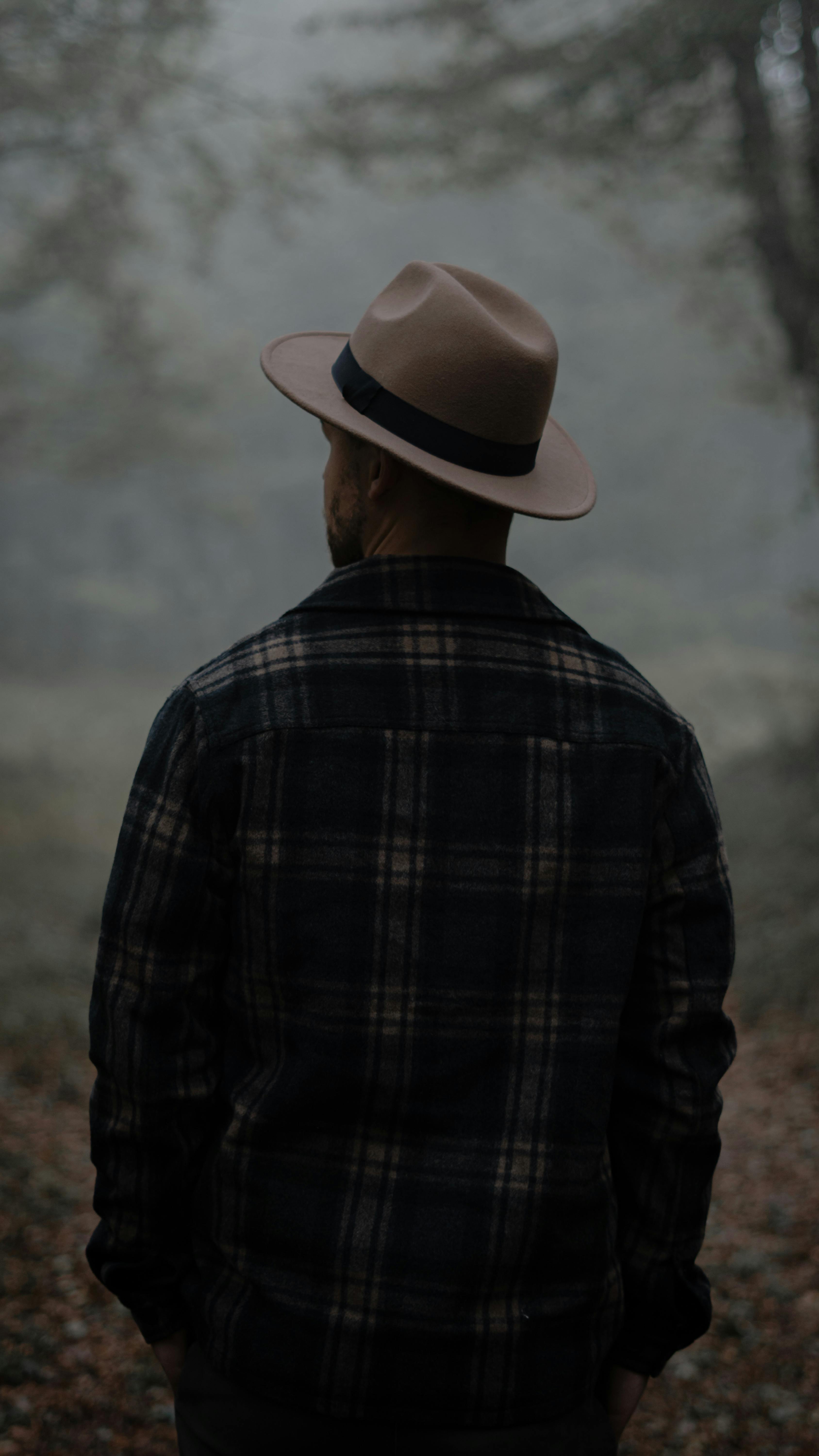 Back view of anonymous male in hat standing in woods with green trees and fallen leaves in misty weather in autumn day