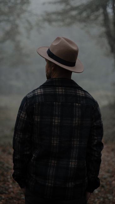 Back view of anonymous male in hat standing in woods with green trees and fallen leaves in misty weather in autumn day