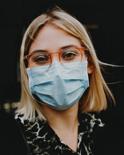 Calm female in eyeglasses wearing protective mask looking at camera while standing on street against blurred background during coronavirus pandemic