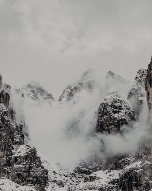 Rough rocky cliffs with steep slopes covered with snow and mist against overcast sky in gloomy weather on cold winter day