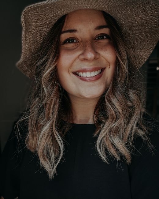 Content female with wavy hair wearing black outfit and stylish hat smiling and looking at camera while standing in room