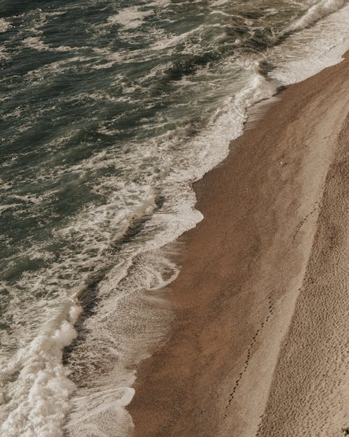 Drone view of rough wavy water of sea with foam rolling on sandy coast with trails in tropical country in nature