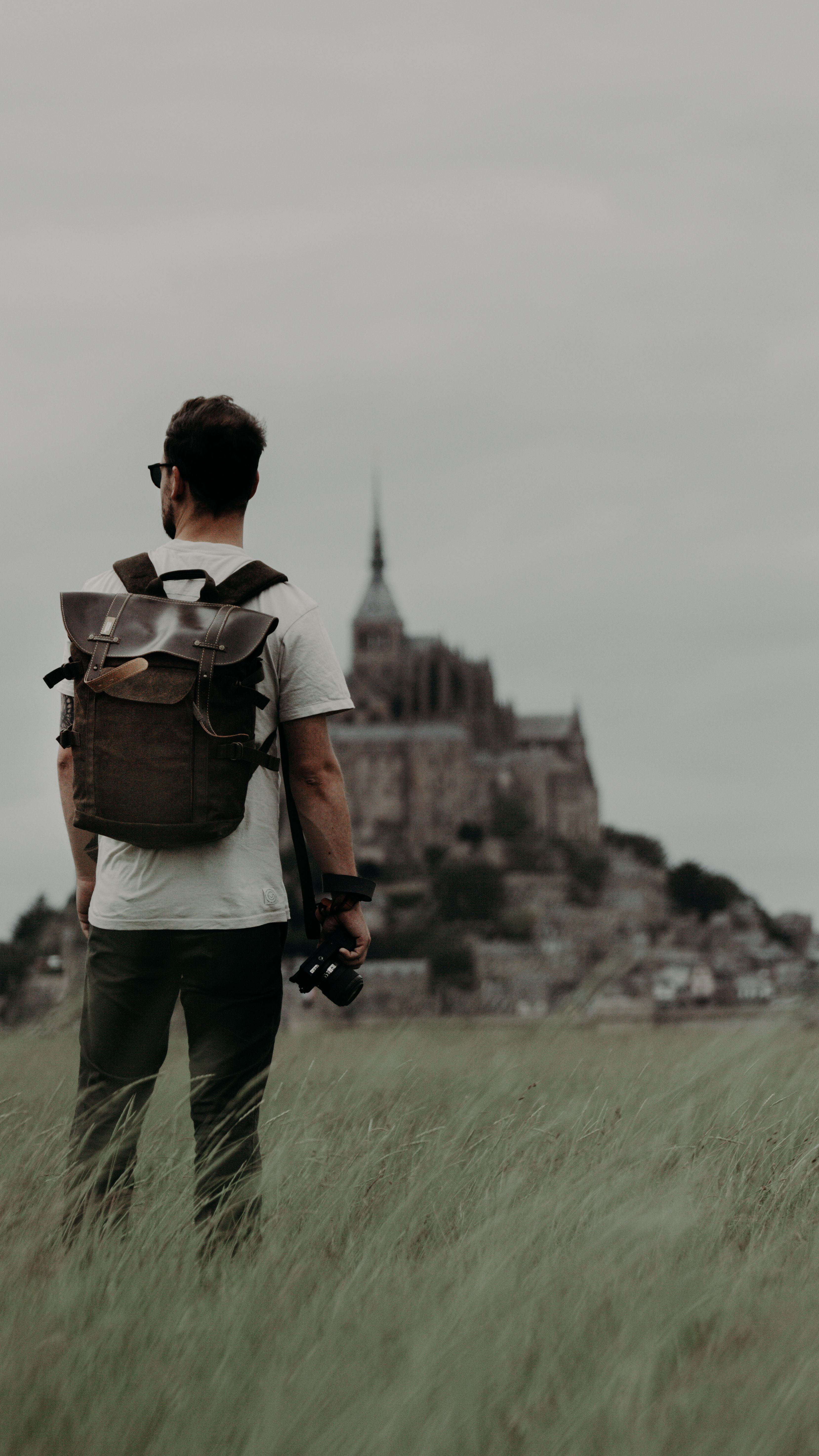 Back view of unrecognizable male traveler with rucksack and photo camera in hand standing in grassy field against aged historic buildings