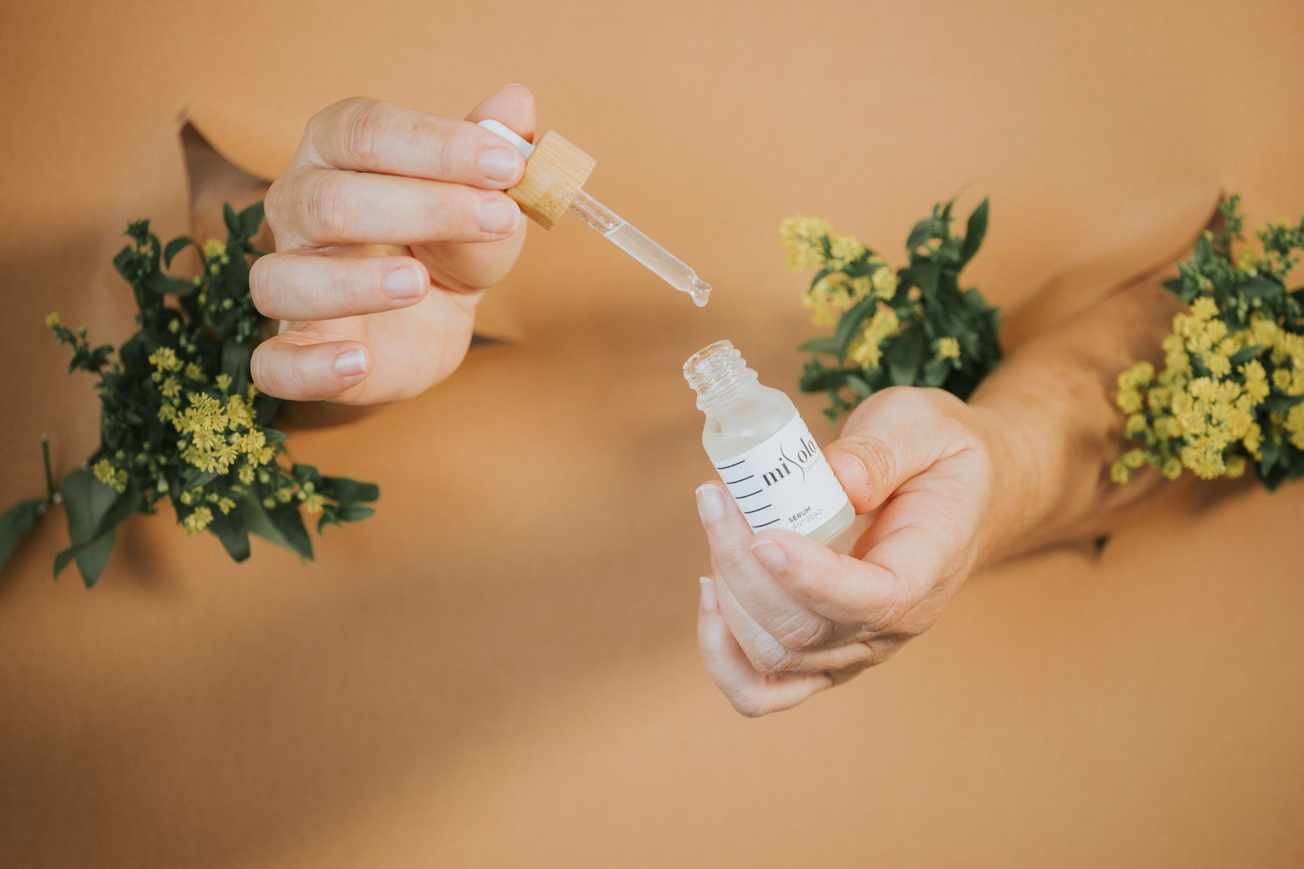 Close-up of hands holding a pipette over a serum bottle with greenery background, promoting natural skincare.