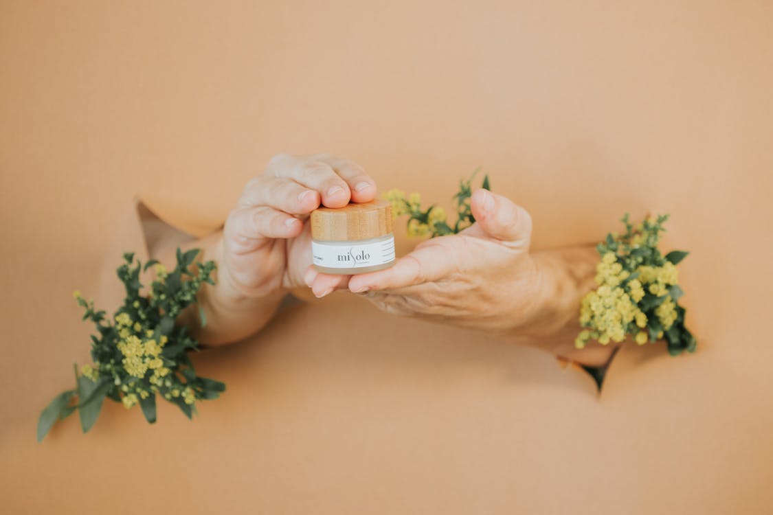 Hands Breaking Through the Paper Wall Holding a Container with Face Cream 