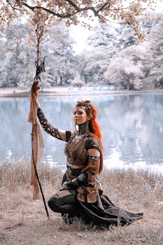Red-haired woman in warrior costume posing with staff by a serene lake.