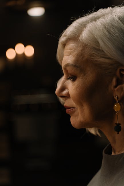 Close-up profile of a senior woman with stylish earrings and white hair against a dark backdrop.