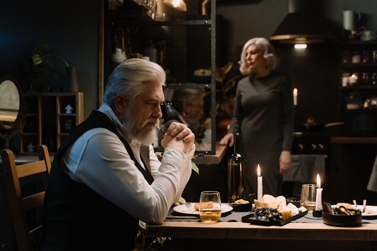 Elderly Man Sitting At The Table With Food And Candles And Woman Standing In The Background 