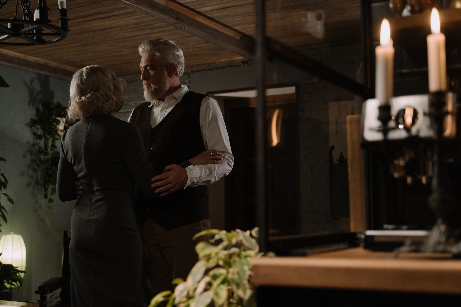 An elegant elderly couple shares a warm embrace in a cozy, candle-lit room.